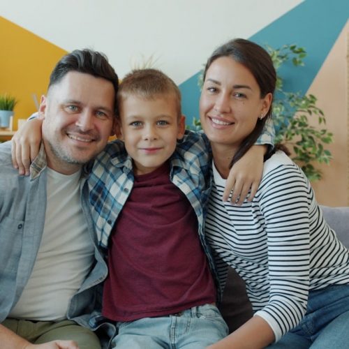 portrait of happy family mother, father and son smiling looking at camera sitting on couch in modern apartment. People and lifestyle concept.
