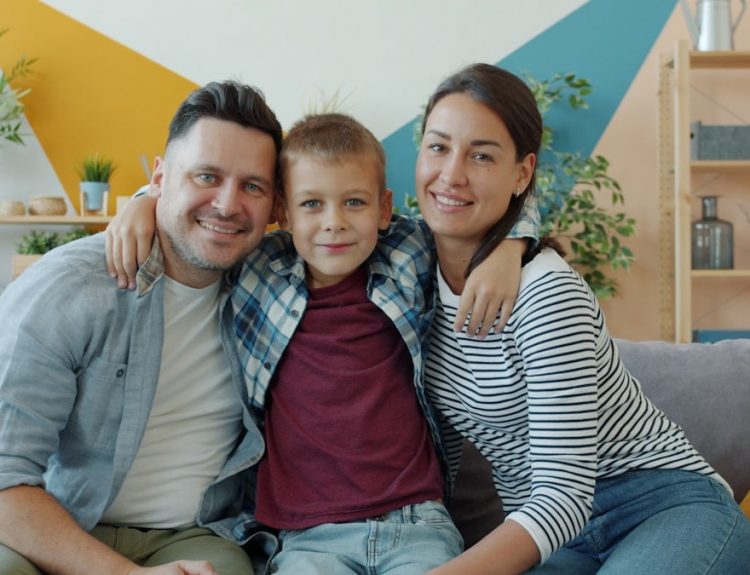 portrait of happy family mother, father and son smiling looking at camera sitting on couch in modern apartment. People and lifestyle concept.