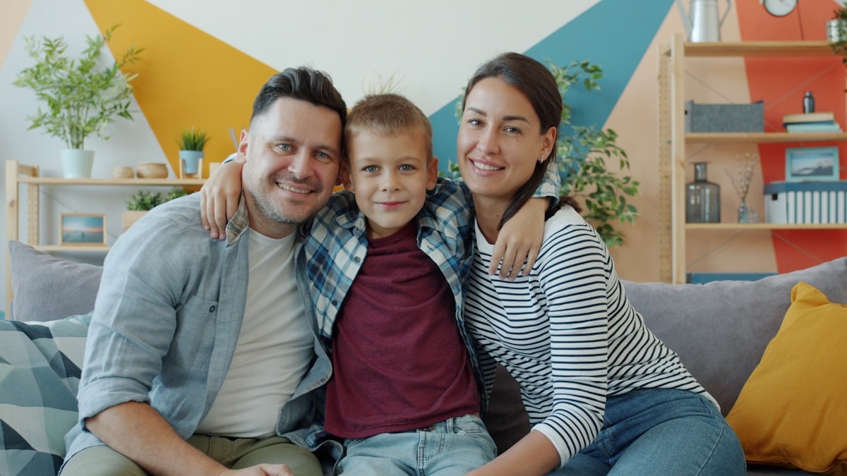 portrait of happy family mother, father and son smiling looking at camera sitting on couch in modern apartment. People and lifestyle concept.